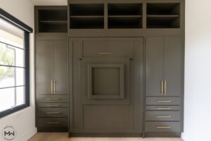 Custom-built dark olive queen Murphy bed with gold hardware, integrated into a full wall storage unit. The design includes open upper shelving, side cabinets with drawers, and a unique fold-out desk in the center of the bed frame. The room features light hardwood flooring, a large window with black trim, and a modern, sophisticated aesthetic.