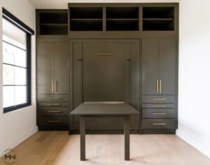 Custom-built dark olive queen Murphy bed with gold hardware, integrated into a full wall storage unit. The design includes open upper shelving, side cabinets with drawers, and a unique fold-out desk in the center of the bed frame.