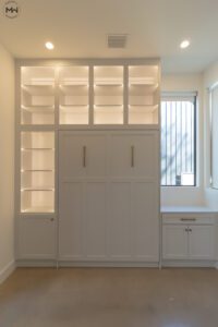 A modern home office featuring a built-in white Murphy bed with gold handles, surrounded by lighted open shelves.