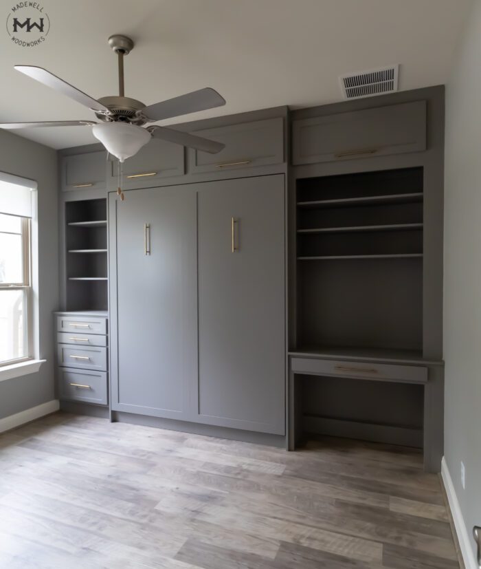 Custom-built gray queen Murphy bed with gold handles, integrated into a multifunctional wall unit featuring built-in shelving, drawers, overhead cabinets, and a compact desk. The room has light wood flooring, a ceiling fan, and a large window allowing natural light to enhance the modern, space-saving design.