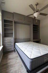 Custom-built gray queen Murphy bed with gold handles, integrated into a multifunctional wall unit featuring built-in shelving, drawers, overhead cabinets, and a compact desk. Features a open bed with a 12" Lull Premium mattress.