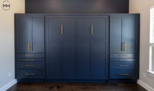 A custom-built navy blue king Murphy bed with shaker-style paneling, integrated into a wall-to-wall storage unit with gold handles. The design includes side cabinets with drawers and doors for additional storage. The dark wood flooring and accent wall enhance the sophisticated, modern aesthetic.
