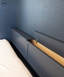 Close-up of a custom-built navy blue pillow storage headboard with a lift-up compartment, featuring a soft-close hinge and natural wood interior. The headboard is part of a modern bedroom design with textured navy walls and a neatly made bed with a patterned white mattress cover.