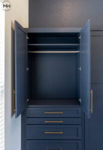 Open navy blue built-in wardrobe cabinet with gold handles, featuring a hanging rod, upper storage shelf, and multiple drawers below. The modern design integrates seamlessly with the surrounding cabinetry in a sophisticated, space-saving layout.