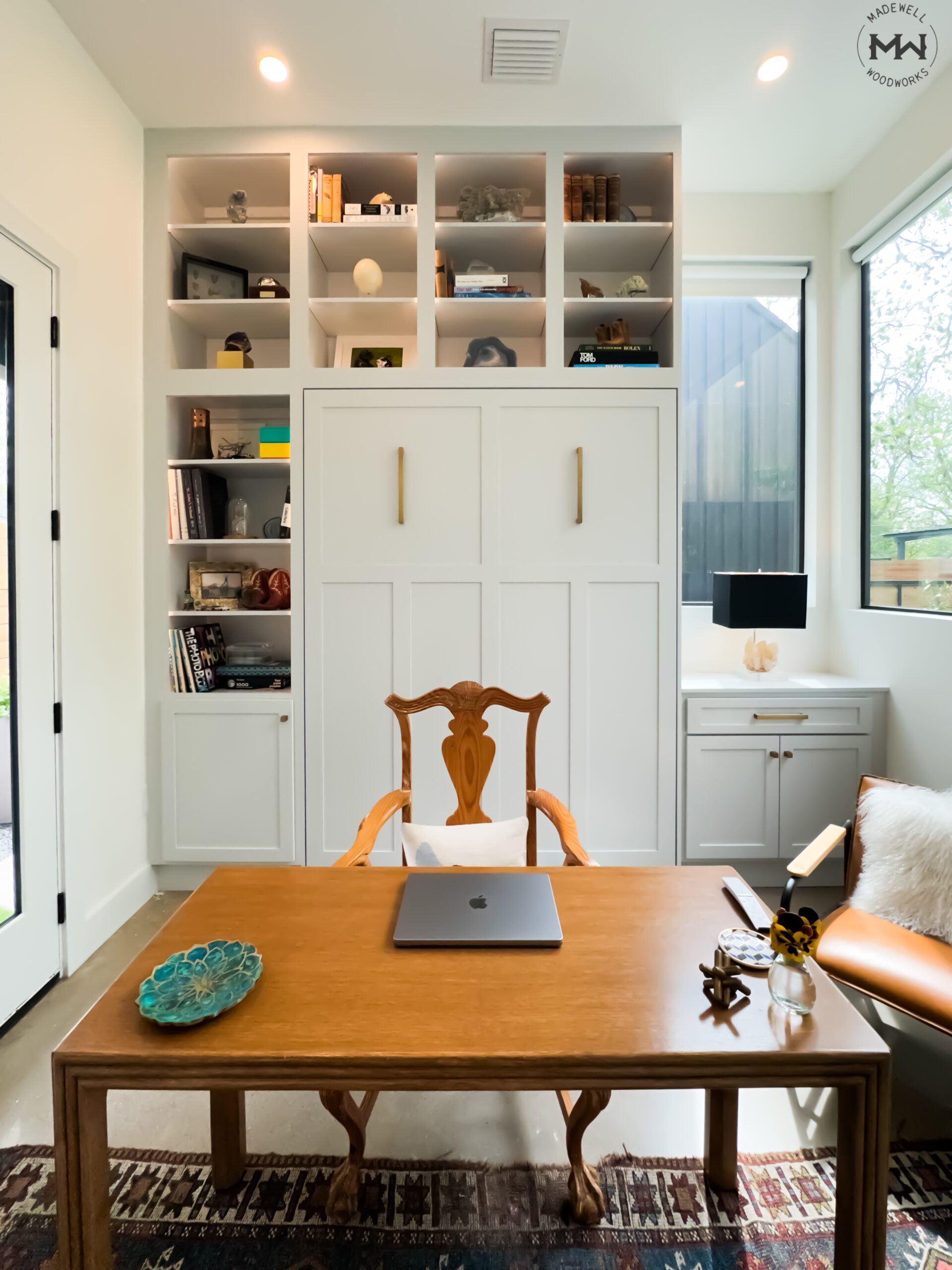 A modern home office featuring a built-in white Murphy bed with gold handles, surrounded by open shelves displaying books, decorative items, and storage boxes. A wooden desk with a classic carved chair sits in the foreground, with a laptop placed on top. To the right, a cozy leather chair with a white fur pillow is next to a small side table holding a vase with flowers. Large windows allow natural light to fill the space, highlighting the functional and stylish design