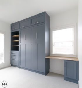A custom-built, navy blue wall unit featuring a Murphy bed with craftsman doors, open shelving with wood accents, and multiple drawers with sleek silver handles. To the right, a built-in desk with a light wood surface is positioned under a window with white blinds. The unit combines storage and workspace in a modern, functional design.