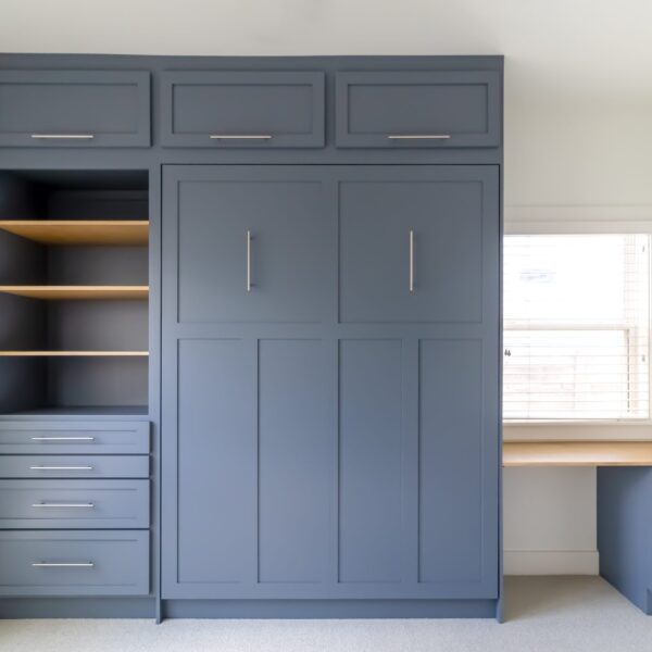 A custom-built, navy blue wall unit featuring a Murphy bed with craftsman doors, open shelving with wood accents, and multiple drawers with sleek silver handles. To the right, a built-in desk with a light wood surface is positioned under a window with white blinds. The unit combines storage and workspace in a modern, functional design.