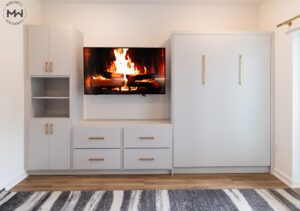 A modern built-in wall unit with light gray cabinetry and gold handles. A flat-screen TV is mounted in the center, displaying an image of a burning fireplace. To the left, there are open shelves and cabinets for storage. To the right, a large Murphy bed is integrated into the design.