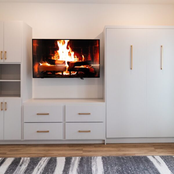 A modern built-in wall unit with light gray cabinetry and gold handles. A flat-screen TV is mounted in the center, displaying an image of a burning fireplace. To the left, there are open shelves and cabinets for storage. To the right, a large Murphy bed is integrated into the design.