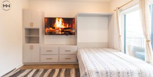 A modern built-in wall unit with light gray cabinetry and gold handles. A flat-screen TV is mounted in the center, displaying an image of a burning fireplace. To the left, there are open shelves and cabinets for storage. To the right, a large open queen Murphy bed is integrated into the design.
