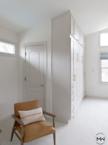 Side view of a custom-built light gray Murphy bed seamlessly integrated into a floor-to-ceiling storage unit with gold handles. The design includes multiple drawers, cabinets, and overhead storage. The room is bright with natural light coming from large windows with soft blue Roman shades. A mid-century modern leather armchair sits in the corner, adding warmth to the neutral space.