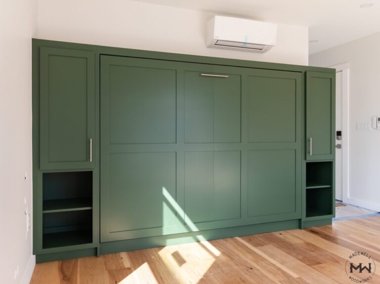 Modern hunter green Murphy bed with custom cabinetry and integrated shelving in a bright room with hardwood floors.