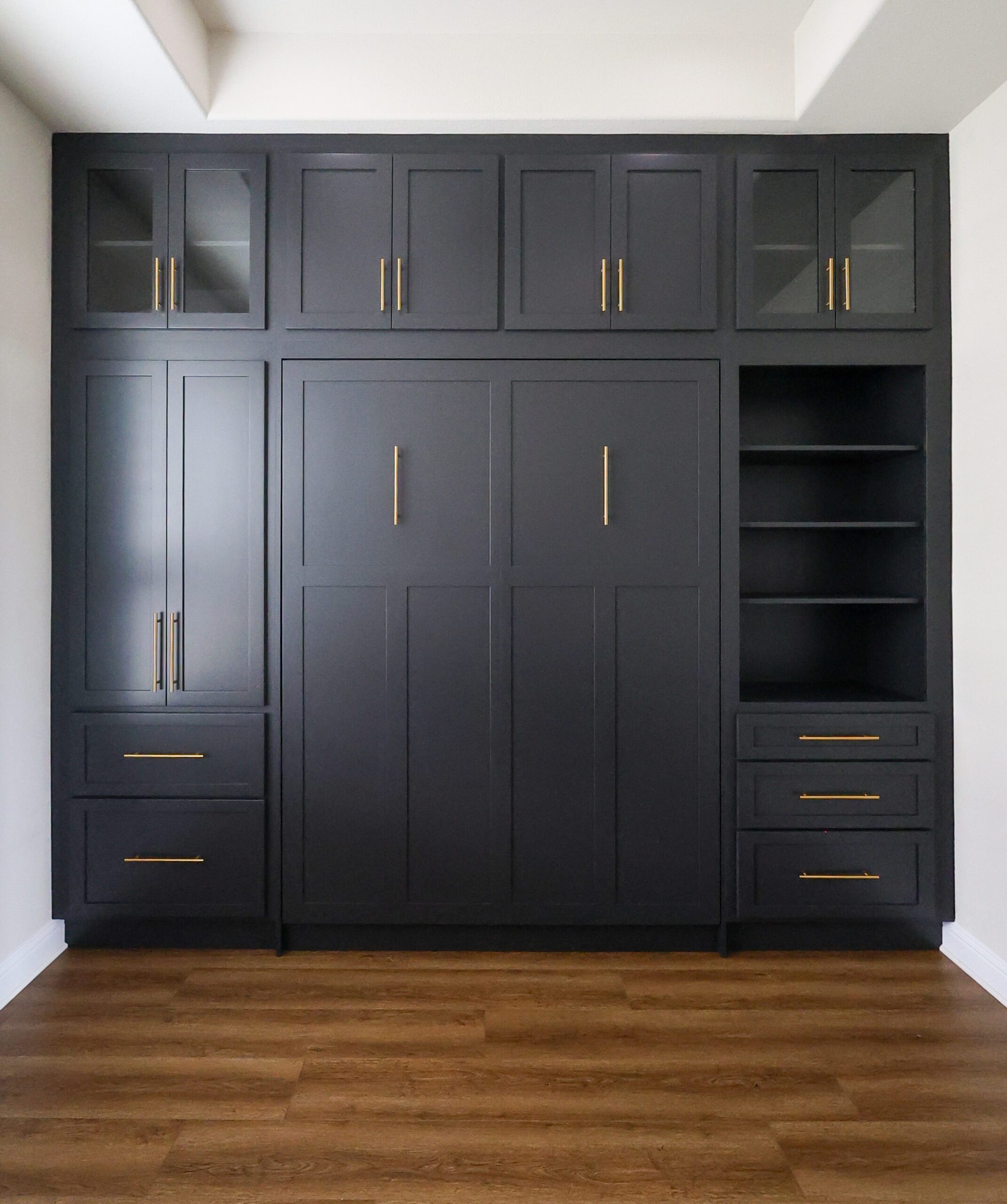 Built-in black cabinets with glass-front doors and gold handles, spanning wall, with open shelving on the right and drawers below.